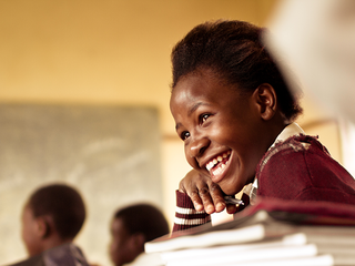 A Happy young South African girl (from the Xhosa tribe) works on her studies and jokes with her friends at at an old worn desk in a class room in the Transkei region of rural South Africa.